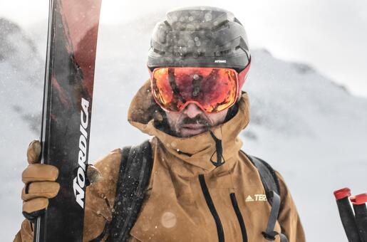 Skier with helmet, ski goggles and skis in hand in front of snowy mountain backdrop